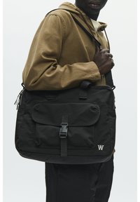 Borsa a tracolla nera in nylon con patta frontale e fibbia, dotata di tasca laterale con cerniera e dettaglio con logo. Indossata sulla spalla.