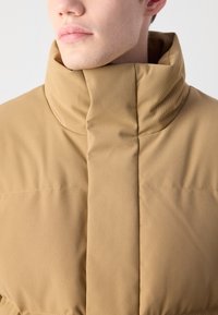 Béžová vypchávkovaná bunda má vysoký golier, textúrovanú tkaninu a elegantný strih. Materiál sa javí ako odolný a nepriepustný na vrásky.