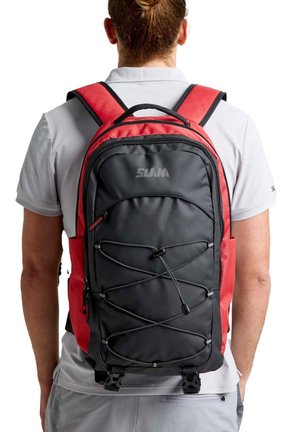 Una persona vista desde atrás lleva una mochila negra y roja con cordones elásticos en la parte delantera y varios compartimentos con cremallera.