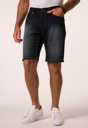 Mann trägt dunkle Jeansshorts, weiße Sneakers und ein weißes Hemd, mit einer Hand in der Tasche und einem Armband am Handgelenk.