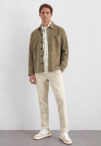 Olivgrüne Button-Down-Jacke mit zwei Fronttaschen, getragen über einem weißen Hemd mit grünen Streifen, kombiniert mit hellbeigen Hosen und weißen Sneakers.
