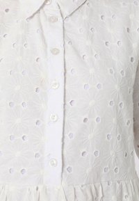White embroidered blouse featuring flower patterns with cut-out detailing, button closure, and a ruffled hem. Soft, textured fabric.