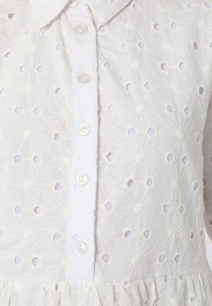 Blusa bianca ricamata con motivi floreali, dettaglio a taglio, chiusura con bottoni e orlo arricciato. Tessuto morbido e testurizzato.