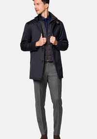 Navy płaszcz zewnętrzny z dwiema guzikami z przodu, pod nim teksturowana szara kamizelka, szare eleganckie spodnie i brązowe buty. Czyste linie i nowoczesny krój.