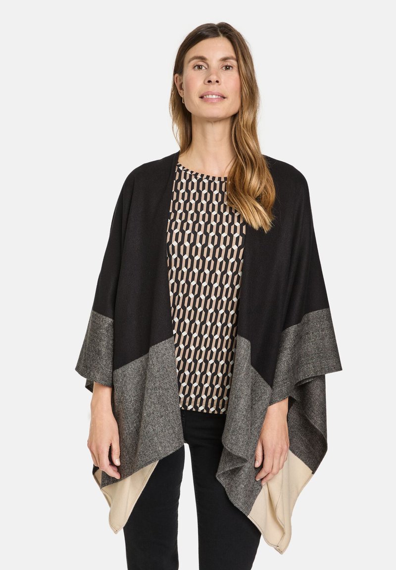 Sort og grå lagdelt poncho med et draperet design, der har kontrasterende teksturer og en beige kant, båret over en mønstret bluse.