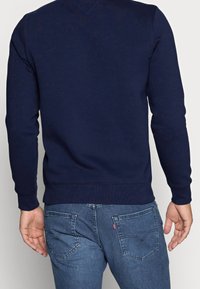 Mörkblå sweatshirt med långa ärmar, rund halsringning och ribbade ärmar; bärs över ljusblå jeans med klassisk bakfickadesign.