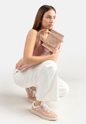 Donna accovacciata con pantaloni bianchi, top rosa e sneakers rosa, che tiene vicino al volto una piccola borsa rosa con cinturini con fibbia.