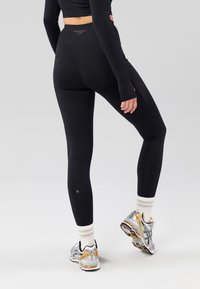 Legging noir de sport avec une ceinture haute et une texture lisse, présentant des coutures discrètes et un petit logo près de la cheville.