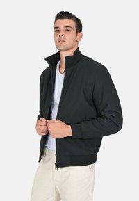 Giacca bomber nera con colletto alto, chiusura con zip, tasche laterali e polsini e orlo a costine. Indossata sopra una maglietta bianca.