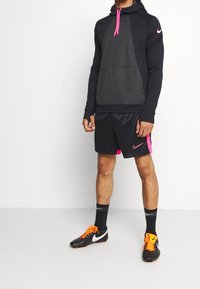 Mężczyzna ubrany w czarną i ciemnoszaro-skalną bluzę Nike, czarne szorty z różowymi akcentami, czarne skarpetki Nike oraz czarne korki z pomarańczowymi sznurowadłami i białymi logotypami.