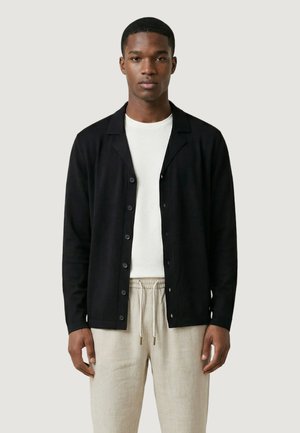 Muž v černém propínacím svetru přes bílé tričko a béžových kalhotách se šňůrkou v pase, stojící na neutrálním pozadí.