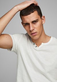 Witte T-shirt met korte mouwen en een subtiele textuur, voorzien van een V-hals en twee zwarte knopen onder de halslijn.