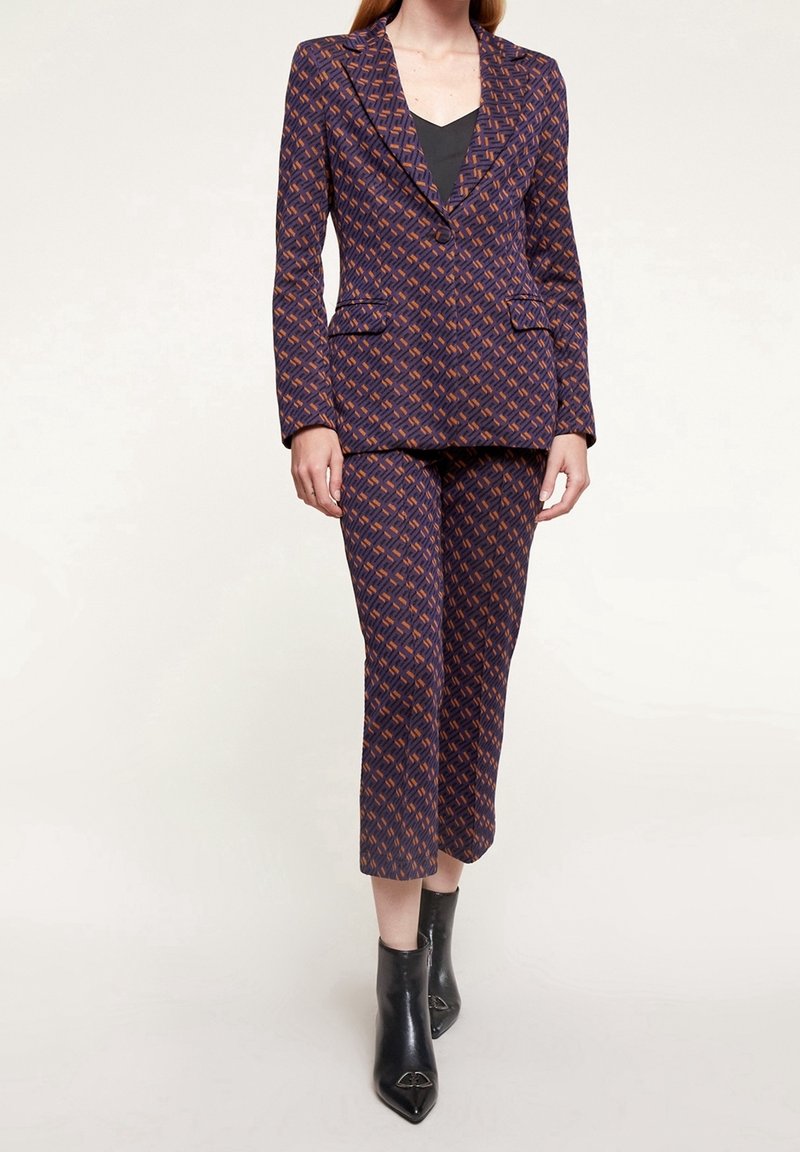 Námořnický oblek s abstraktním oranžovým vzorem, obsahující přiléhavý blejzr s klopami a odpovídající zúžené kalhoty; zkombinováno s černými kotníkovými botami.