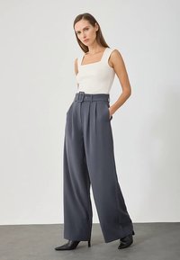 Žena u bijelom bez rukava i širokim hlačama tamnosive boje visokog struka s odgovarajućim remenom, stoji s rukama u džepovima.