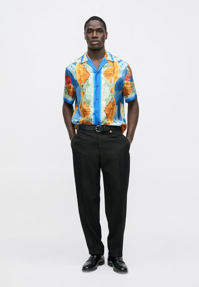 Κοντομάνικο πουκάμισο με έγχρωμο σχέδιο χάρτη, που περιλαμβάνει αποχρώσεις μπλε, πορτοκαλί και πράσινο. Συνδυασμένο με μαύρο παντελόνι και λαμπερά μαύρα παπούτσια.