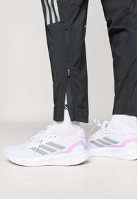 Λευκά αθλητικά παπούτσια με γκρι ρίγες και ροζ λεπτομέρειες, σε συνδυασμό με μαύρο στενό παντελόνι φόρμας που διαθέτει φερμουάρ στον αστράγαλο.