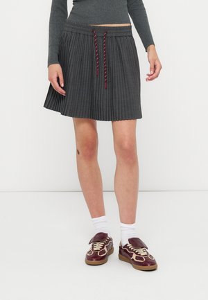 Person iført en mørk plisseret nederdel med røde snore, hvide sokker og bordeaux sneakers står mod en ensfarvet hvid baggrund.