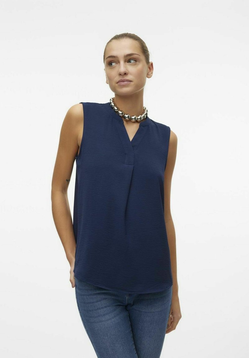 Blouse sans manches bleu marine avec un col en V et un tissu texturé. Associée à un jean ajusté et un collier métallique massif.