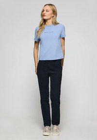 T-shirt en coton bleu clair avec "Polo Club" imprimé au centre, associé à un pantalon marine foncé et des baskets beige. Le modèle se tient debout.