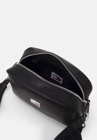 Bolso bandolera de piel negra con una cremallera plateada. En su interior cuenta con un suave forro de tela y una etiqueta de marca con acentos de color. Incluye una correa ajustable.