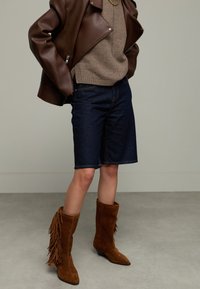 Braune Lederjacke, beiger Pullover, dunkle Denim-Shorts und braune Wildleder-Stiefel bis zum Knie mit Fransen-Details. Strukturierter und schichtartiger Look.