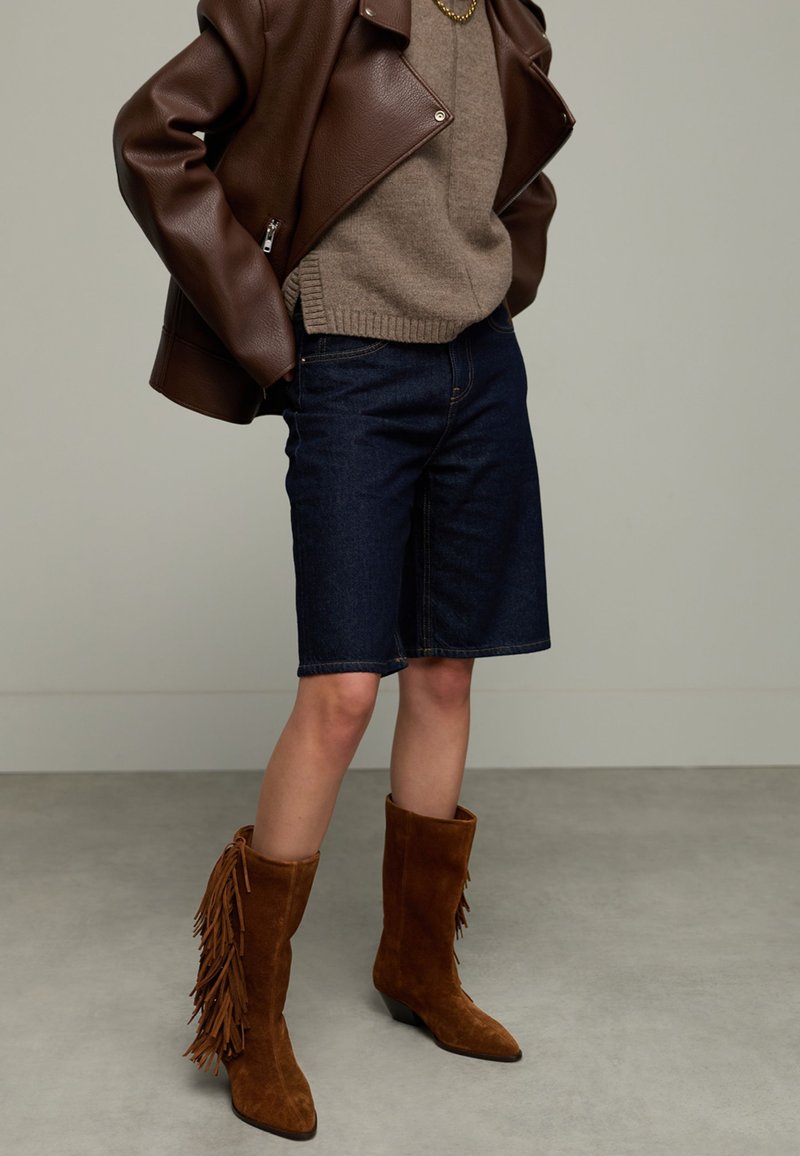 Braune Lederjacke, beiger Pullover, dunkle Denim-Shorts und braune Wildleder-Stiefel bis zum Knie mit Fransen-Details. Strukturierter und schichtartiger Look.