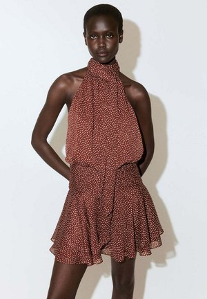 Femme debout face à l'avant, vêtue d'une robe marron sans manches à col montant avec des pois blancs et une jupe à volants.