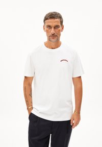 Weißes T-Shirt mit kurzen Ärmeln aus Baumwolle. Fässt einen kleinen roten Logo-Schriftzug auf der linken Brust. Sauberes, einfaches Design mit Rundhalsausschnitt.