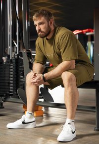 Maglietta atletica verde oliva e pantaloni corti. Sneakers bianche con accenti neri. Tracker fitness nero al polso. Sfondo neutro da palestra.