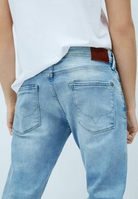 Vaaleansiniset farkut, joissa on sileä pinta, viisi taskua ja ruskea nahkapaikka vyötäröllä. Niissä on myös kulutettuja yksityiskohtia.