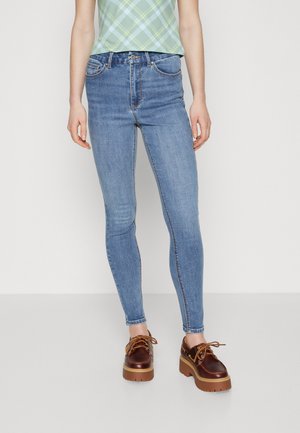 Light blue skinny jeans with five-pocket design, belt loops, and subtle stitching details, paired with brown platform shoes and a green plaid top.
