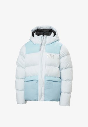 Jeune fille souriante, portant une veste d'hiver rembourrée bleu clair et blanc avec une capuche et un jean bleu, debout devant un fond blanc.