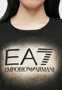 Fekete póló elülső grafikával, melyen vastag fekete és enyhén arany színű "EA7" felirat látható, alatta az "EMPORIO ARMANI" felirat és egy szárny logó.