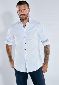 Weißes Hemd mit knöpfbarem Kragen und hochgerollten Ärmeln, verziert mit einem gemusterten Besatz. Schwarze Knöpfe und eine dezente Textur betonen das Design. Kombiniert mit dunklen Jeans.