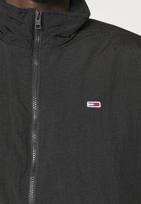 Giacca nera con zip e collare alto, caratterizzata da una consistenza liscia e leggera e da una piccola patch con logo rosso, bianco e blu sul petto.