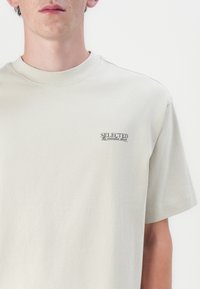 Hellbeige Baumwoll-T-Shirt mit kurzen Ärmeln. Verfügt über ein kleines, schwarzes, gesticktes "SELECTED"-Logo auf der linken Brustseite.