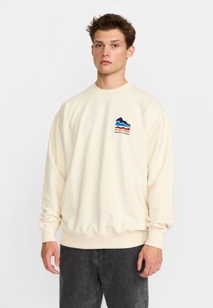 Joven con el pelo corto y castaño que lleva una sudadera crema con un logo de ola colorido y vaqueros negros, de pie frente a un fondo liso.