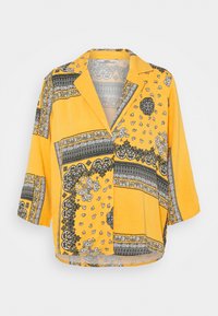 Blusa gialla con vestibilità rilassata, caratterizzata da motivi paisley e geometrici in bianco e nero. Ha un colletto a punte e maniche a tre quarti.