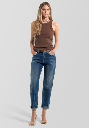 Mujer con pelo largo y rubio, que lleva una camiseta marrón sin mangas, vaqueros azules remangados, un cinturón y sandalias de tacón beige, de pie con las manos en los bolsillos.
