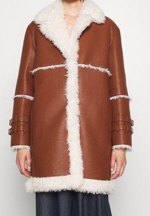 Brun læderjakke i brystlængde med hvidt shearling-for langs krave, forstykke, manchetter og kant, båret over mørke bukser.