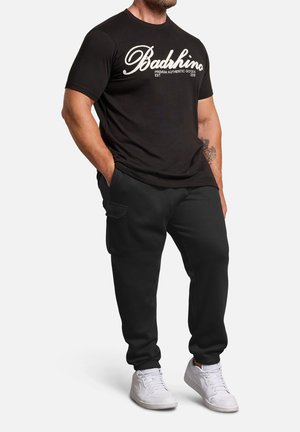 Mann trägt schwarzes BadRhino-T-Shirt, schwarze Jogginghose mit Seitentasche und weiße Sneaker, steht mit den Händen in den Taschen.