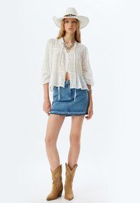 Blusa blanca de botones con textura y anudada al frente, combinada con una falda mini de mezclilla. Botas de vaquero marrones y un sombrero blanco de ala ancha.