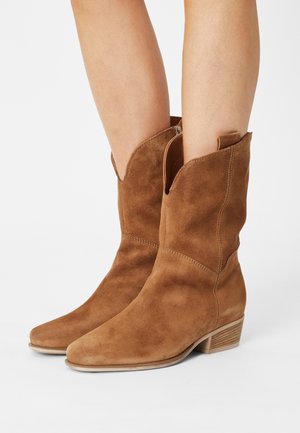Bruine suède enkelboots met een taps toelopende neus en een lage gestapelde hak, voorzien van decoratieve stiksels en zijhandvaten voor gemakkelijk aan- en uittrekken.