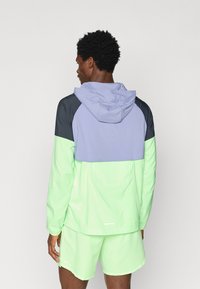 Homme portant une veste à capuche colorée et un short assorti, debout de dos face à un fond uni.