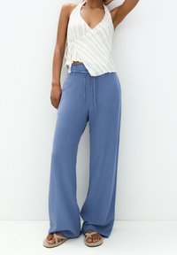 Biała bluzka z paskami, zapinana na dekolt w serek, z detalem wiązania, w połączeniu z jasnoniebieskimi spodniami typu wide-leg i beżowymi sandałami. Minimalistyczny design.