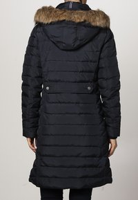 Marineblauwe pufferjas met een gevoerd capuchon van nepbont, gestikte ontwerp, lange lengte en twee drukknoopjes op de rug voor een verstelbare pasvorm.