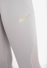 Jasnoszare legginsy z złotym logo Nike oraz teksturowanymi, okrągłymi akcentami na dolnej części nogawek. Wykonane z gładkiego, elastycznego materiału.
