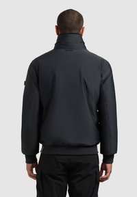 Musta pehmustettu takki, jossa on korkea kaulus, ribattuja hihansuita ja sileä pinta. Takissa on yksinkertainen selkäosa ja minimalistiset yksityiskohdat.