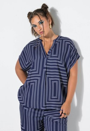 Frau mit geflochtenem Haar, die goldene Creolen, blauen Lidschatten und ein marineblaues Outfit mit weißen geometrischen Linien trägt, posiert vor einem weißen Hintergrund.