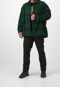 Camicia di flanella a quadri verdi e neri con bottoni, taschino sul petto, T-shirt nera sotto, abbinata a pantaloni neri e sneakers nere.